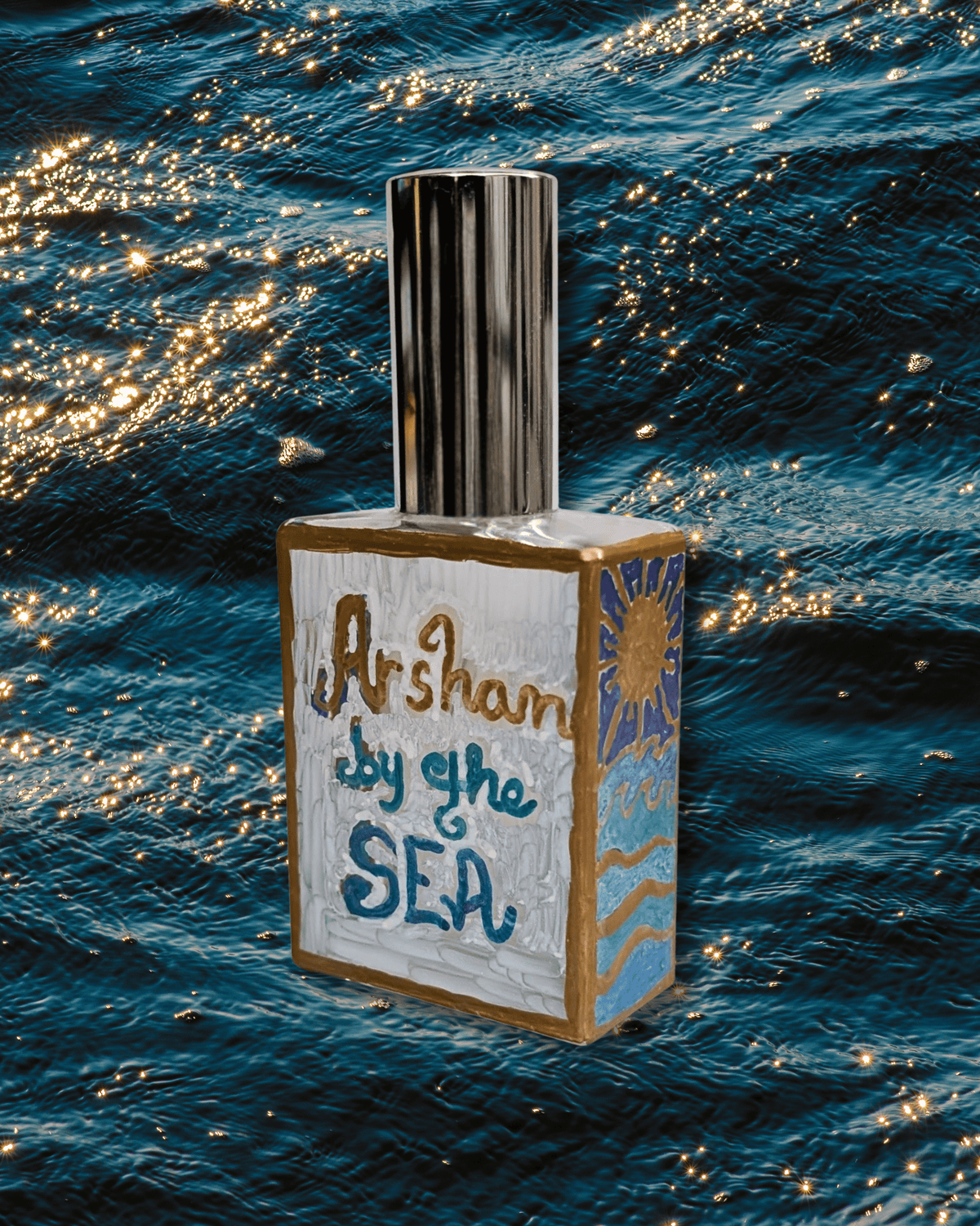 An original hand-painted decorative rectangular bottle with a metallic silver cap, featuring hand-painted artwork on the front and side. The front is painted white with the words 'Arshan by the SEA' in blue and gold lettering, and the side depicts a blue sun and waves with gold accents. The bottle rests over a blue sea glimmering with the golden sun.